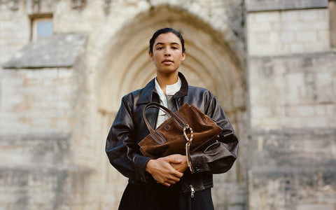 Person wearing a black jacket and skirt, holding a brown suede tote bag in front of a stone building