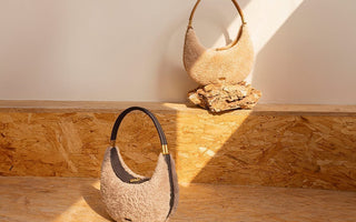 Beige shearling shoulder bag with a unique crescent moon shape and light brown leather handle, displayed on a textured wooden surface