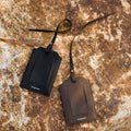 Black and brown leather Songmont luggage tags with clear windows and straps on a rustic rock background.