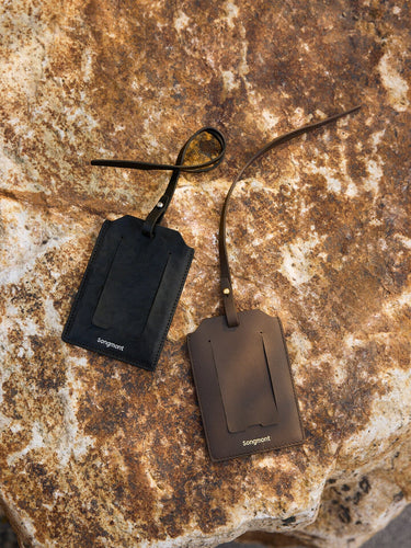 Black and brown leather Songmont luggage tags with clear windows and straps on a rustic rock background.