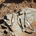 Small Carefree Thread Charm in Sandal color, crafted from wood, leather, and seeds, on a rocky backdrop.