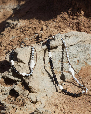 Small Carefree Thread Charm in Sandal color, crafted from wood, leather, and seeds, on a rocky backdrop.