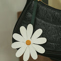 White leather daisy charm with yellow center, attached to a black handbag strap.