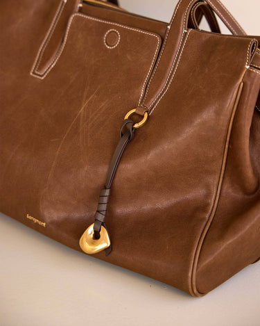 Brown leather Faith Stone Charm with gold hardware, knot detail, and Songmont logo.