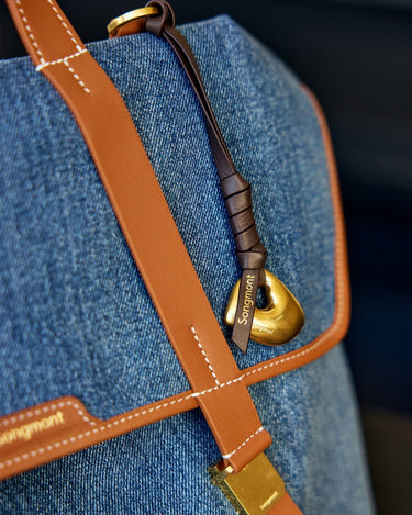 Close-up of Faith Stone Charm in onyx denim with tan leather strap and gold hardware, featuring "Songmont" logo.