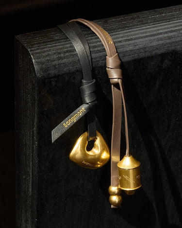 Sandal Faith Stone & Prayer Wheel Charm Set, gold metal charms on black/brown leather straps with Songmont logo.