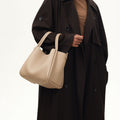 Large Song Bag in cream apricot, pebbled leather with gold feet, held by a model in a brown coat.