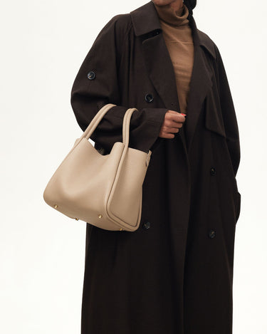 Large Song Bag in cream apricot, pebbled leather with gold feet, held by a model in a brown coat.