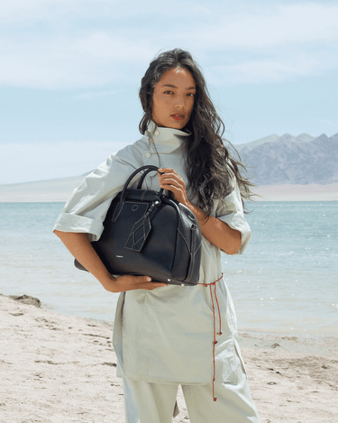 Large black Yore bowling bag made of full-grain Nappa leather with white stitching, held by a woman on a beach.
