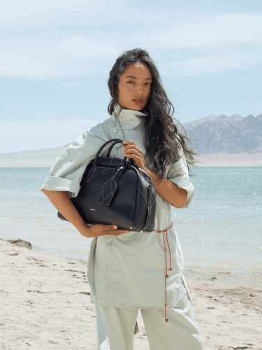 Large black Yore bowling bag made of full-grain Nappa leather with white stitching, held by a woman on a beach.