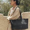 Woman wearing a beige linen suit carrying a large black pebbled leather tote bag with gold hardware.