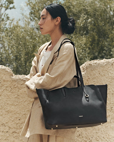 Woman wearing a beige linen suit carrying a large black pebbled leather tote bag with gold hardware.
