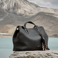 Large Yore Tote Bag in Onyx full-grain leather, with gold hardware and a beaded charm, set against a natural landscape.