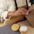 Person applying leather care balm with a cloth to a beige leather bag. Balm in a bamboo tin.