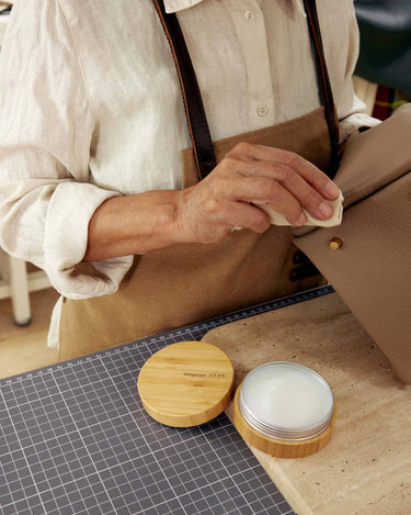 Person applying leather care balm with a cloth to a beige leather bag. Balm in a bamboo tin.