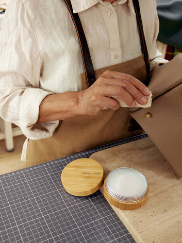 Person applying leather care balm with a cloth to a beige leather bag. Balm in a bamboo tin.
