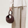 Burgundy Luna Bag made of textured leather with gold hardware, held by a person in a linen shirt and beige trousers.