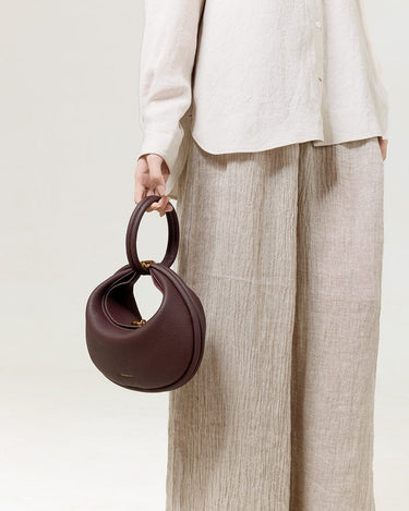 Burgundy Luna Bag made of textured leather with gold hardware, held by a person in a linen shirt and beige trousers.