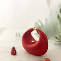Red leather Luna Bag with gold clasp, placed on textured surface with decorative red fruits and a soft, misty background.