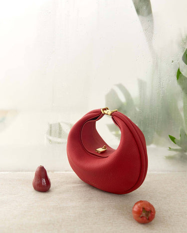 Red leather Luna Bag with gold clasp, placed on textured surface with decorative red fruits and a soft, misty background.