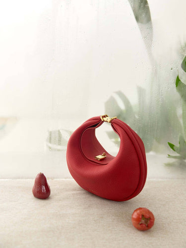 Red leather Luna Bag with gold clasp, placed on textured surface with decorative red fruits and a soft, misty background.