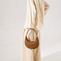 Luna Bag in brown full-grain leather with gold hardware and embossed logo, held by a woman in a linen outfit.