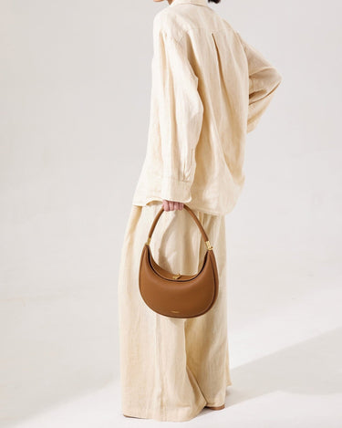 Luna Bag in brown full-grain leather with gold hardware and embossed logo, held by a woman in a linen outfit.