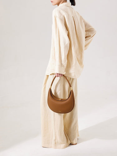 Luna Bag in brown full-grain leather with gold hardware and embossed logo, held by a woman in a linen outfit.