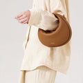 Luna Bag in full-grain leather, brown, crescent-shaped with gold hardware and adjustable strap.