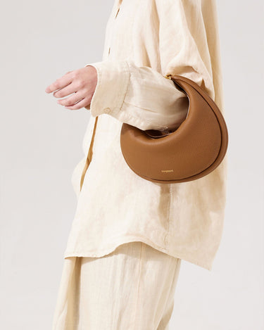 Luna Bag in full-grain leather, brown, crescent-shaped with gold hardware and adjustable strap.