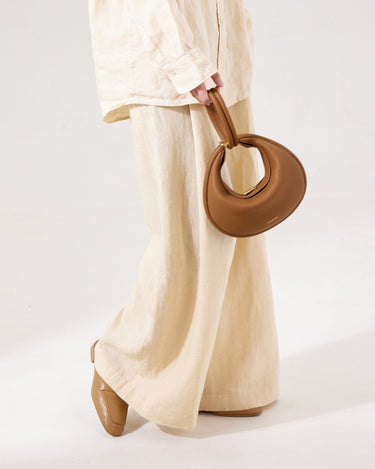 Brown full-grain leather Luna bag with gold clasp, held by person in linen outfit.