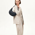 Woman wearing a beige suit and carrying a black Luna Bag on her shoulder, featuring gold hardware and a curved silhouette.