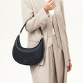A woman wears a tan suit and holds a black pebbled leather Luna Bag with gold hardware.