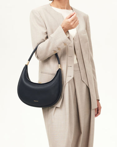 A woman wears a tan suit and holds a black pebbled leather Luna Bag with gold hardware.