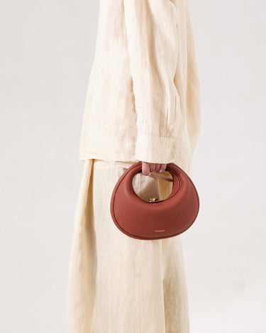 Luna Bag in Oolong: A round, pebble-grain leather handbag with a gold clasp, held by a person in a beige linen outfit.