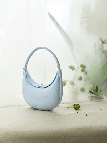 Light blue Luna Bag with pebble-grain leather texture, silver clasp, and curved handle on textured surface.