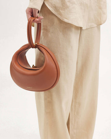 Luna Bag in Eco Caramel vegan leather with a gold clasp, held by a person in beige linen clothing.