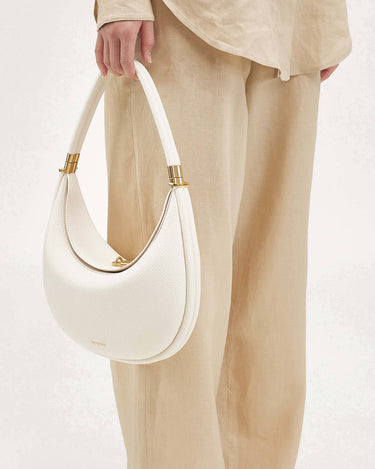 White vegan leather Luna Bag with gold hardware, held by a person wearing beige linen pants and a light-colored top.