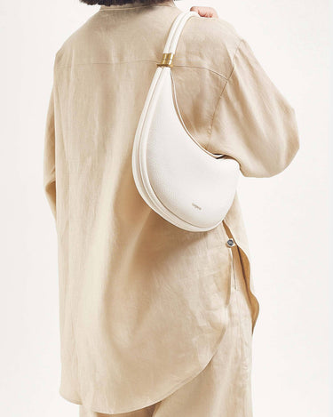 White vegan leather Luna Bag with gold hardware worn on the shoulder by a person in a beige linen outfit.