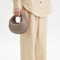 Taupe full-grain leather Luna Bag with gold hardware, shown being held by a person in beige linen clothing.
