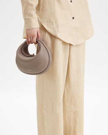 Taupe full-grain leather Luna Bag with gold hardware, shown being held by a person in beige linen clothing.