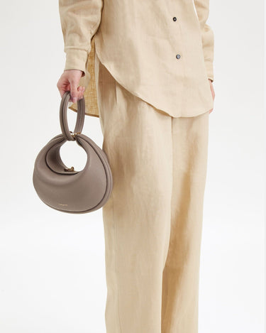 Grey full-grain leather Luna Bag with top handle and gold hardware, held by a person in a linen outfit.