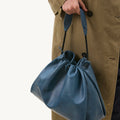 Navy blue leather Medium Gather Bag Set with gathered top, braided straps, held by person in tan trench coat.