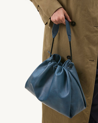 Navy blue leather Medium Gather Bag Set with gathered top, braided straps, held by person in tan trench coat.