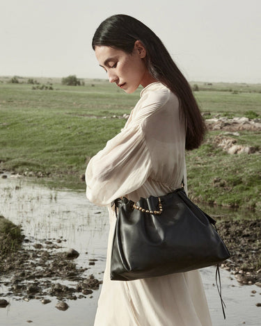Woman in a flowing cream dress carrying an Onyx Medium Gather Bag with gold bead detail