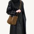 Medium Song Bag in Agarwood brown suede with gold hardware, worn by a person in a black leather coat.