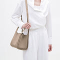 Medium Song Bag in Taupe: A pebbled leather bucket bag worn by a person in a white outfit.