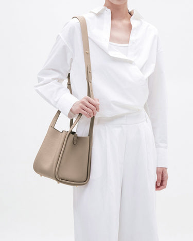 Medium Song Bag in Taupe: A pebbled leather bucket bag worn by a person in a white outfit.