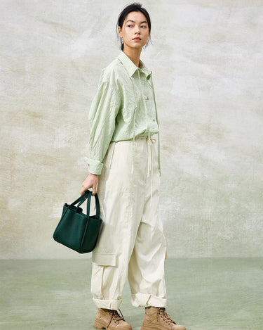 Medium Song Bag in emerald green leather, held by a woman wearing a casual green shirt and off-white cargo pants.