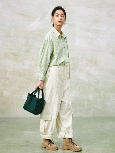 Medium Song Bag in emerald green leather, held by a woman wearing a casual green shirt and off-white cargo pants.